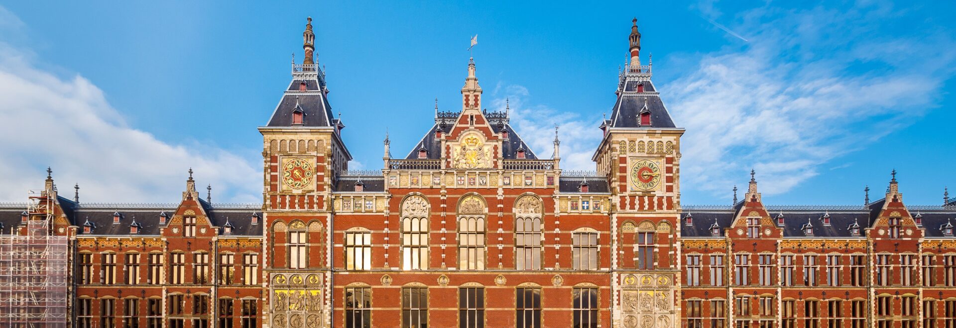 Vooraanzicht station Amsterdam Centraal met het stationsplein. Het steenrode/gouden gebouw ontworpen door Cuypers steekt prachtig af tegen de helder blauwe lucht met lichte sluierbewolking.