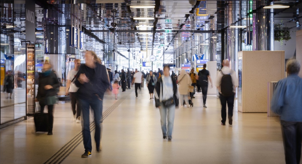 Wide shot van de IJhal op Amsterdam Centraal, met diverse mensen die aan het wandelen zijn. De mensen zijn wazig, alsof ze in beweging zijn.
