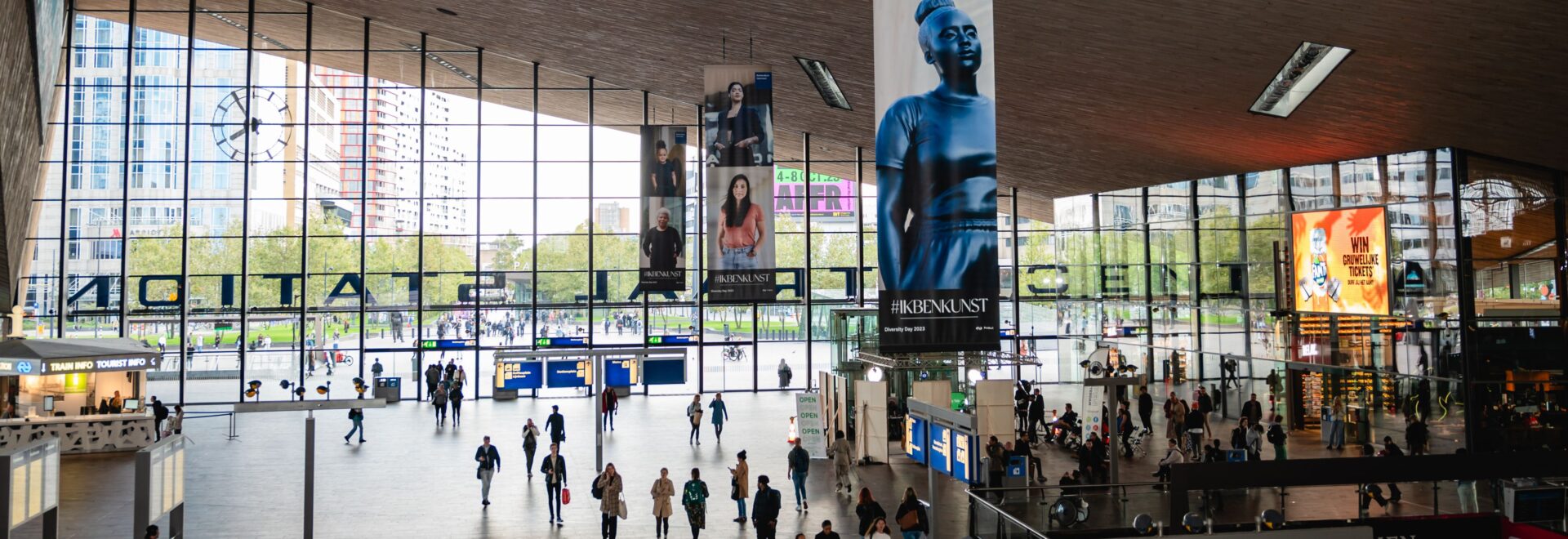 Foto van de stationshal van Rotterdam Centraal met een banner van het beeld 'Moments Contained' met bezoekers van het station er onder als mieren. Foto gemaakt in het kader van Diversity Day 2023 door Michelle Urbiztondo.