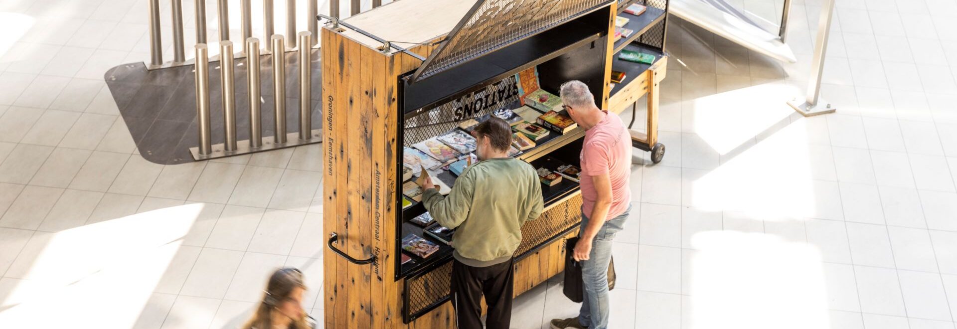 Station Book Swap in the hall of Utrecht Central Station. You see two people in front of the StationsBoekWissel browsing through the available books.