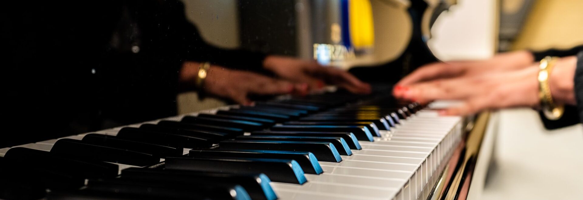 de toetsen van de stationspiano aan de linkerzijde van het beeld. Rechts op de toetsen zie je onscherp handen die de piano op de hoogste toetsen bespelen. Rode nagellak en een gouden armband om de arm. Reflectie van de persoon die piano speelt is te zien in het gelakte zwarte hout van de vleugel.