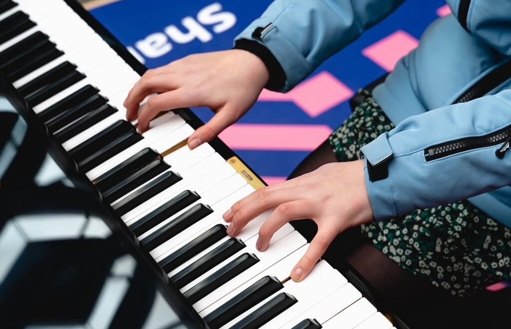 De handen van pianiste Sera op de toetsen van de Stationspiano op Den Haag Centraal. In de zwart glimmende deksel van de vleugel zie je de reflectie van het Wiebertjesdak.